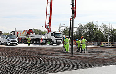 Betong pumpas ut på en stor yta med armeringsjärn. Tre gulklädda arbetare styr röret som pumpar ut betongen. I bakgrunden två vita betongbilar.