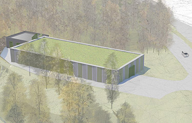 En grå avlång byggnad med grönt tak i ett skogsparti. Intill byggnaden går en väg.