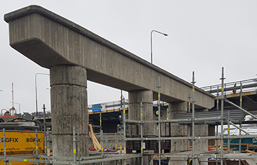 Fyra betongpelare med en ovanpåliggande betongkonstruktion. Runtomkring syns byggställningar och i bakgrunden en bro.