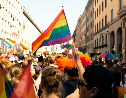 Människor firar på en Pride-parad med regnbågsflaggor på en solig gata. Foto: William Fonteneau/Unsplash