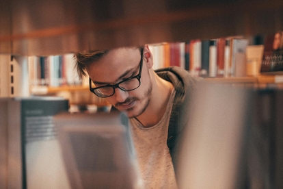 Person med glasögon läser bok i bibliotek. Foto: MalteLuk/Pexels