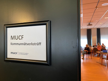 Skylt på dörr med texten "MUCF Kommunnätverksträff" och en grupp människor som sitter i ett mötesrum i bakgrunden. Foto: Eva Blässar, MUCF