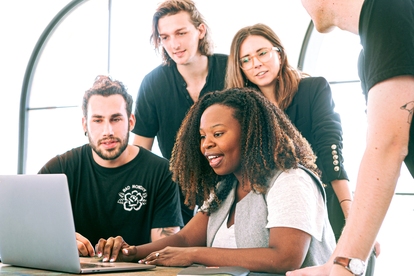 Grupp av unga personer diskuterar runt en laptop i ett ljust kontor. Foto: Canva Studio/Pexels