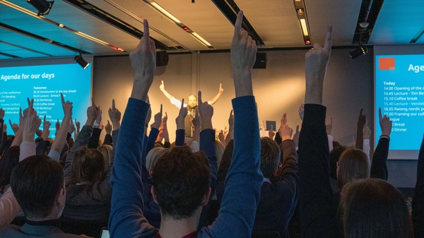 Personer på en konferens räcker upp händerna i en interaktiv session med en talare i bakgrunden. Foto: MUCF.