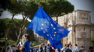 Ung person viftar med en EU-flagga i en samling av unga människor utomhus framför en historisk byggnad. Foto: Antoine Schibler/Unsplash.
