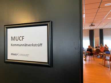 Skylt med texten "MUCF Kommunätsverksträff" på vägg vid mötesrum med människor i bakgrunden. Foto: MUCF