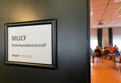 Skylt med texten 'MUCF Kommunnätsverksträff' på en vägg med utsikt mot ett mötesrum där människor sitter och samtalar. Foto: MUCF.