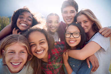 Grupp av leende ungdomar som kramar varandra utomhus i soligt väder.