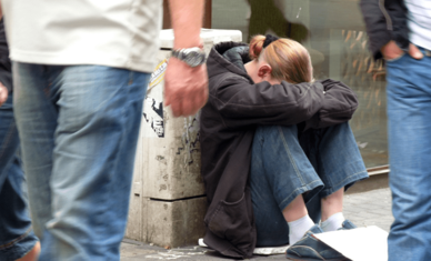 Ung person sittande på gatan med huvudet i händerna medan människor passerar förbi.