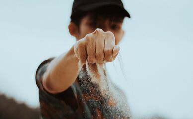 Närbild på en hand som låter sand rinna mellan fingrarna, suddig person i bakgrunden.