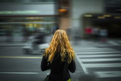 Person med långt hår sedd bakifrån vid ett övergångsställe i stad med rörelseoskärpa i trafiken.