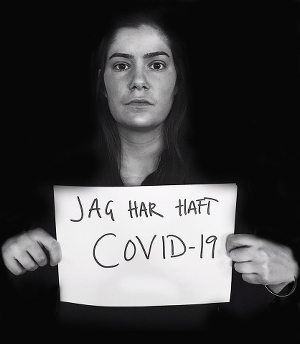 Ett svartvitt fotografi av en kvinna. Kvinnan ser in i kameran och håller upp ett vitt papper. På pappret står det med svart text "Jag har haft Covid-19".