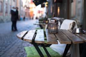 Ett cafébord står på trottoaren utanför ett fik. På bordet står en ljuslykta. Bredvid bordet står en stol med en dyna och en filt. Fler cafébord syns längre bort.