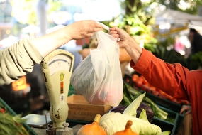 En arm sträcks över en disk för att lämna över en påse med grönsaker. På andra sidan disken sträcks en annan arm fram för att ta emot påsen.