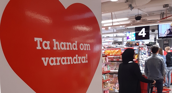 Rött hjärta med texten Ta hand om varandra! fotograferat framför en kassakö i en butik.