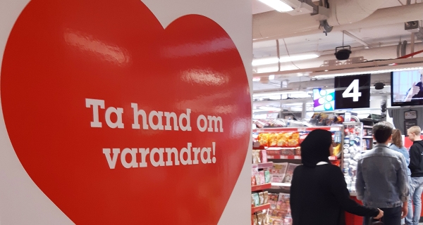 Rött hjärta med texten Ta hand om varandra! fotograferat framför en kassakö i en butik.