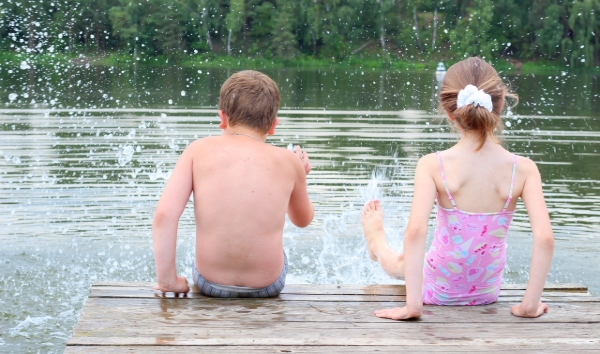 Två badklädda barn sitter på en brygga och plaskar med benen i vattnet så att det stänker.