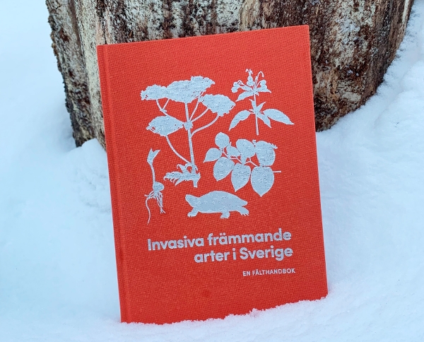 Boken "Invasiva främmande arter i Sverige" står ute i snön lutad mot en trädstam.