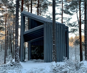Öppen gråmålad träbyggnad med kraftiga bjälkar i snöig tallskog.