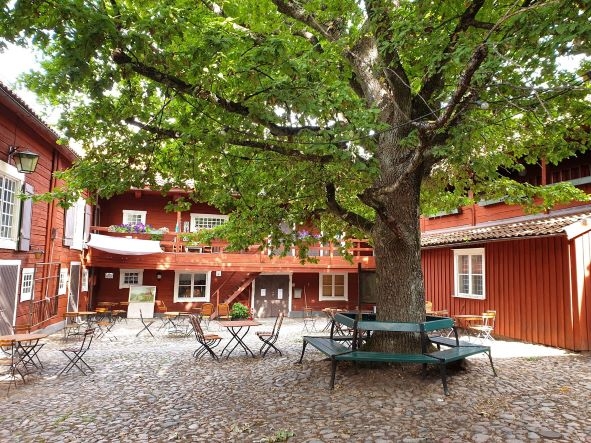Gammal röd träbyggnad som omsluter en kullerstensbelagd innegård med stolar och bord och ett stort grönt träd i mitten med en bänk runt stammen.