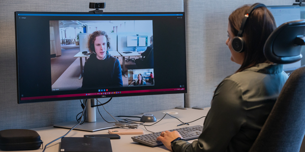 Person sitter vid skrivbord och deltar i videomöte med person på stor datorskärm