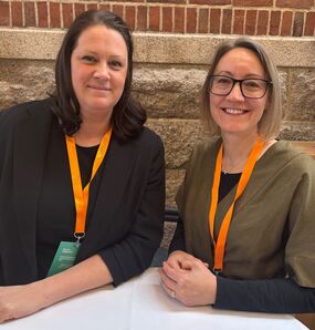 Två kvinnor med orange ID-band står vid ett bord framför en tegelvägg.