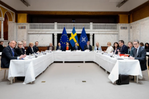 Delegationer sitter runt ett U-format konferensbord med svenska och NATO-flaggor i bakgrunden.