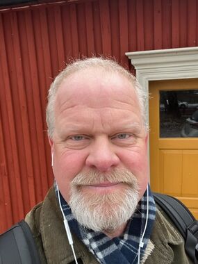 Närbild på leende man med vitt skägg och hörlurar framför röd träbyggnad med gul dörr
