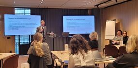 Person som håller presentation för åhörare i mötesrum med bildspel på skärmar. 