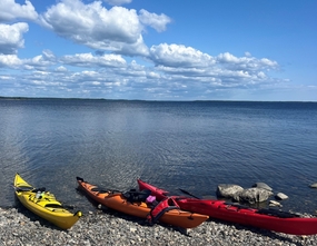 Tre kajaker på klapperstensstrand vid lugnt vatten under blå himmel med moln