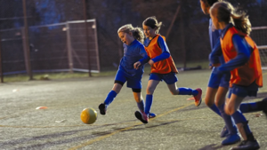 Barn som spelar fotboll på upplyst konstgräsplan under kvällsträning.