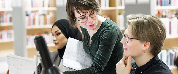 Tre studenter som diskuterar vid ett bord i ett bibliotek.