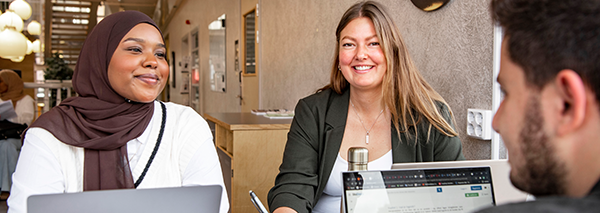 Tre kollegor i ett mötesrum, en man och två kvinnor, varav en bär hijab, samtalar och ler.