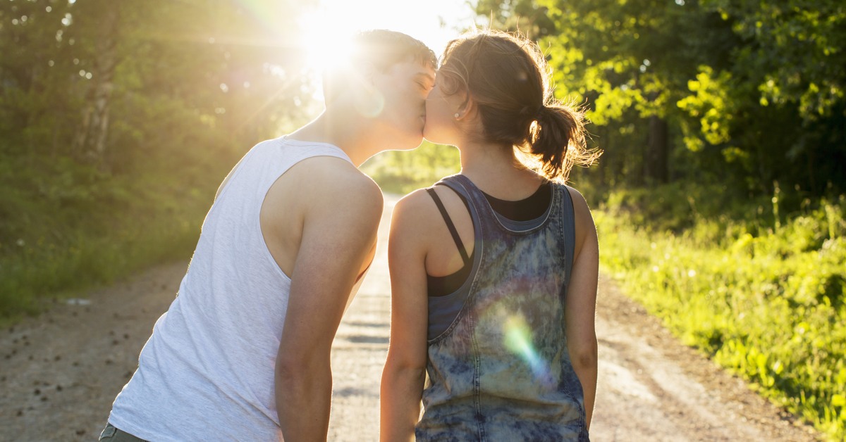 Ett ungt par kysser varandra på en grusväg i motljus.