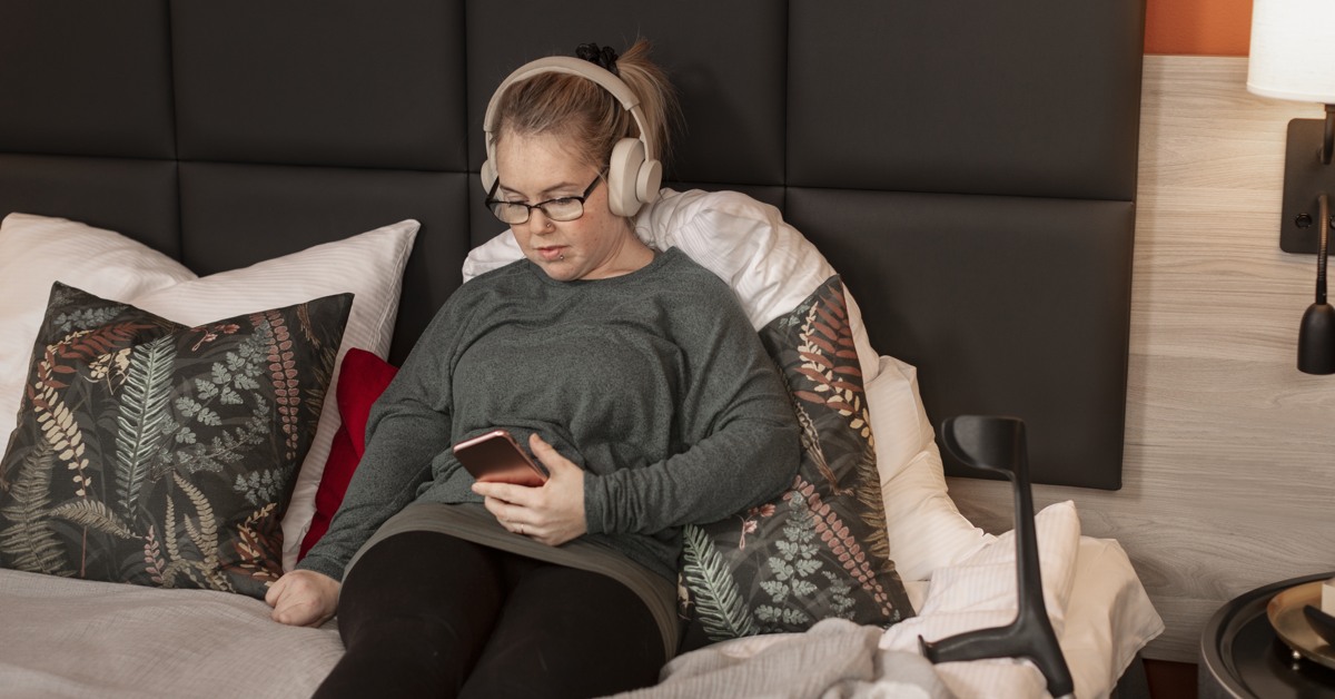 Kvinna ligger på säng med hörlurar och mobiltelefon.