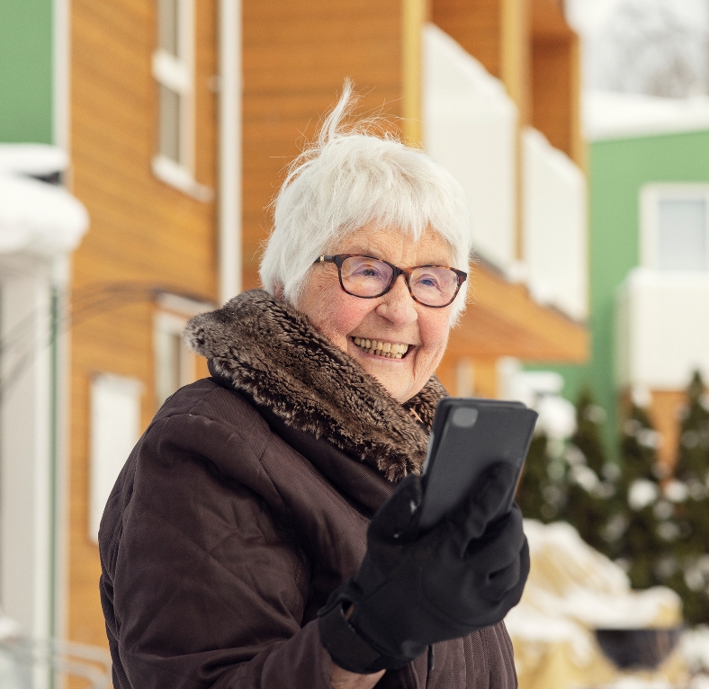 Leende äldre kvinna med glasögon håller i en smartphone utomhus på vintern.