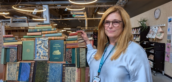 Kvinna med glasögon står bredvid ett stort stapel inbundna böcker i en bibliotekslokal.