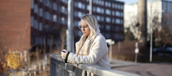 Kvinna i ljus jacka som lutar sig mot ett räcke och tittar på sin mobil.