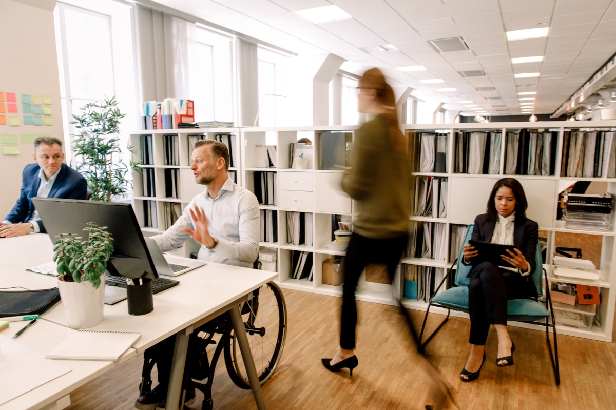 Bilden föreställer ett kontor. En man i rullstol diskuterar med en manlig kollega och två kvinnor syns i bakgrunden.