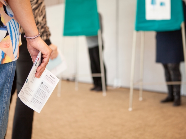 Foto på hans som håller i röstsedel. I suddig förgrund syns två bås där två personer står och röstar. 