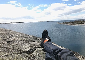 Foto som visar benen och skorna på person som sitter på klipporna på Bullerö och ser ut mot havet och horisonten. Foto: Länsstyrelsen.