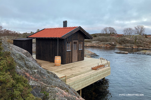 Brun bastustuga med orange tegeltak på en nybyggd brygga i en skärgårdsvik, sen höst. 