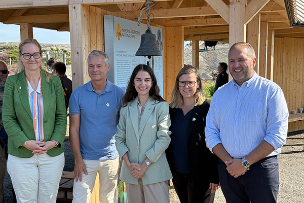 Gruppbild på fyra vuxna människor. Bakom dem ett skärmtak och en informationstavla samt en skeppsklocka.amför ett skärmtak vid en informationstavla.