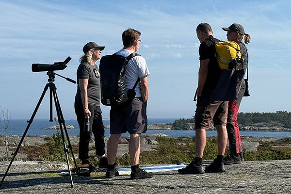 Människor vid en kikare uppe på ett berg en sommardag, hav i bakgrunden.