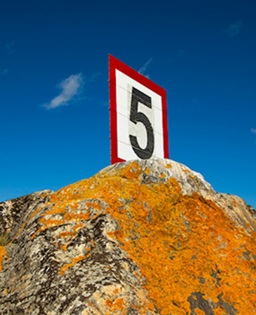 Skylt ute på en klippa utomhus, 5 knop. Blå himmel, sol, gul lav. Foto: Mostphotos.