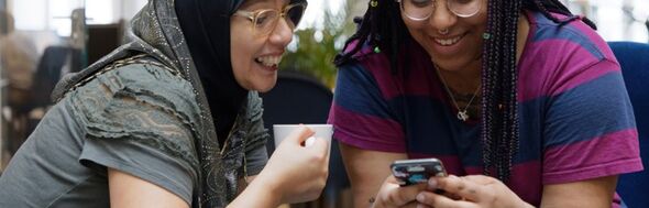 Två glada kvinnor tittar på en mobiltelefon och skrattar tillsammans.