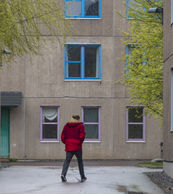 En person i röd jacka står på gården framför en grå lägenhetsbyggnad med blå fönsterkarmar