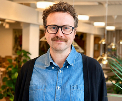 Leende man med glasögon och mustasch i ett ljust, modernt kontorsutrymme.