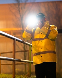 En man från ett vaktbolag står vid en byggnad och lyser med en ficklampa mot kameran.