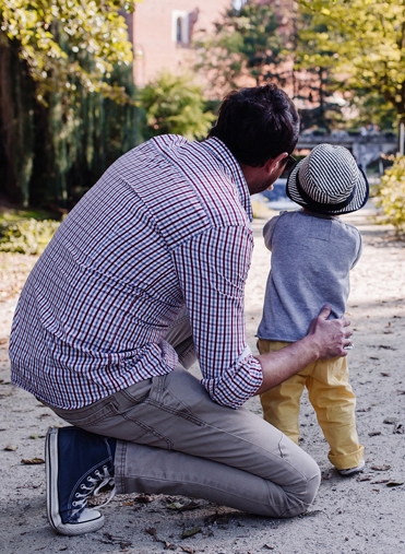 En pappa sitter på huk och håller om ett litet barn, han pekar på något som de tittar på.